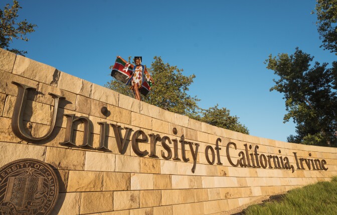 A woman holding a Kenyan flag behind her back stands on top of a wall bearing large brass-colored letters reading "University of California, Irvine" with the UC seal. She is in a celebratory stance. 