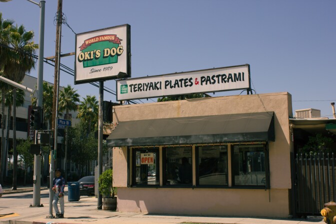 The outside of a small light brown building located on a small street corner. The building contains four identical squared black-framed windows with a slightly faded black awning above. Above the building, it reads Teriyaki Plates & Pastrami in black lettering. Next to the building on the left is a stand-alone sign that reads World Famous Oki's Dog Since 1979. In front of the sign, a dark-skinned man leans against a traffic light pole. 