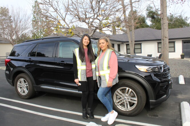 The mental health crisis response workers stand in front of a black SUV. The wear reflective safety vests and are looking towards the camera. 