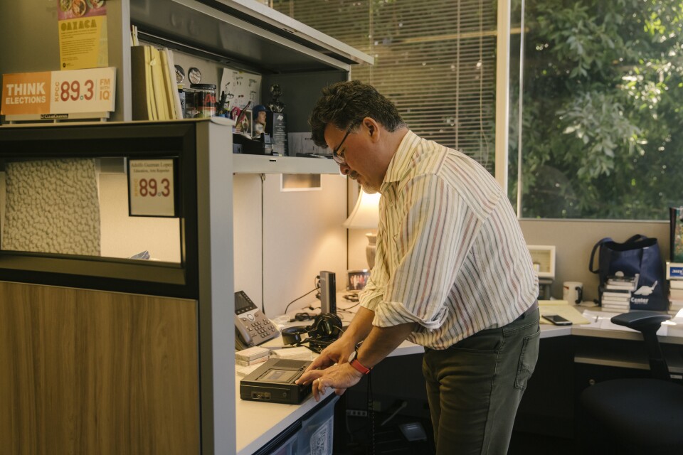 A man in gray slacks and a striped shirt stands over a desk listening to a tape.