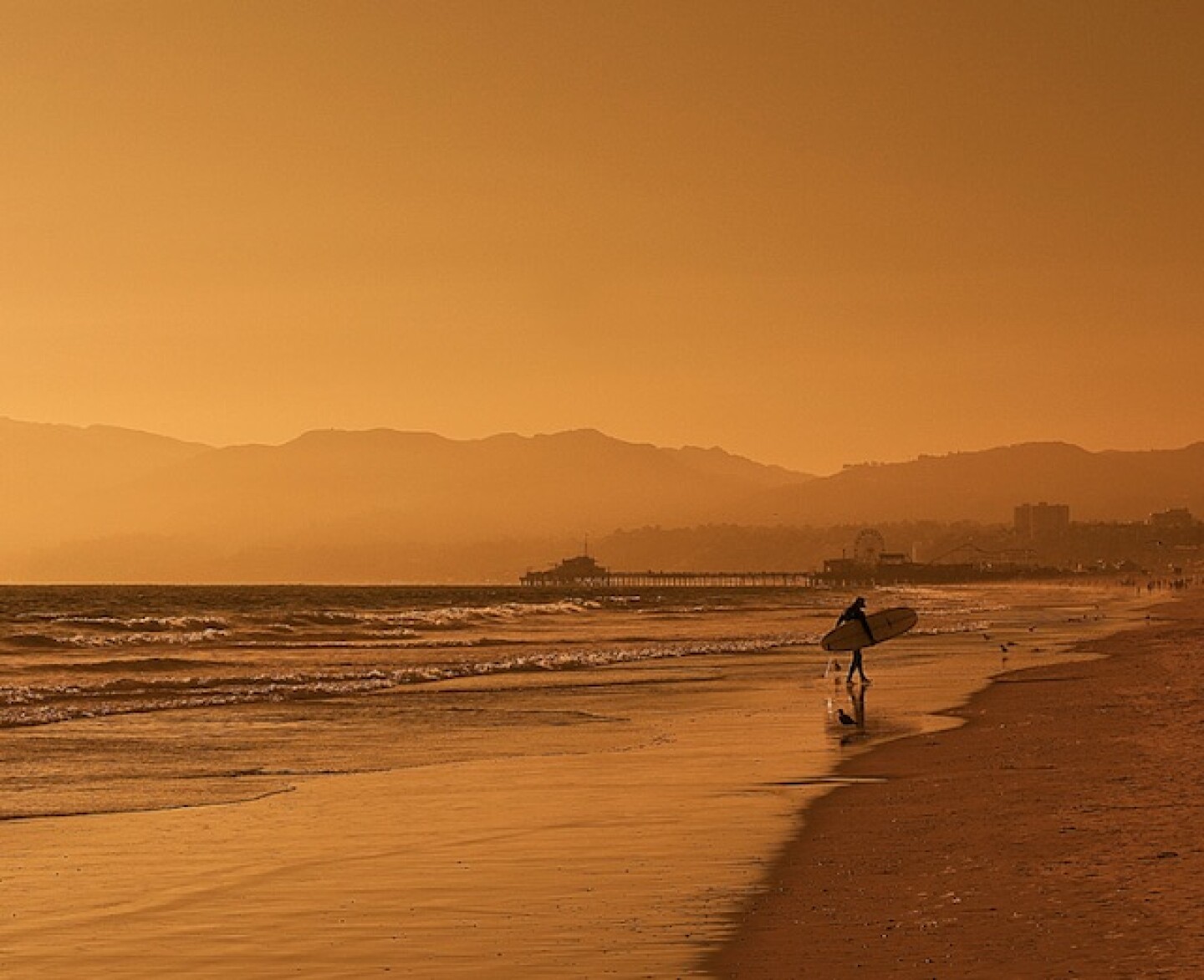 A surfer carries a surfboard along the shoreline on a beach enshrouded in an orange haze.