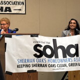 A woman with brown skin and short, light brown hair is behind a lectern. She is wearing a bright blue suit and wearing glasses. To the right of the image, a woman with brown skin and long dark brown hair is behind a separate lectern and microphone. She is wearing a light gray suit. 