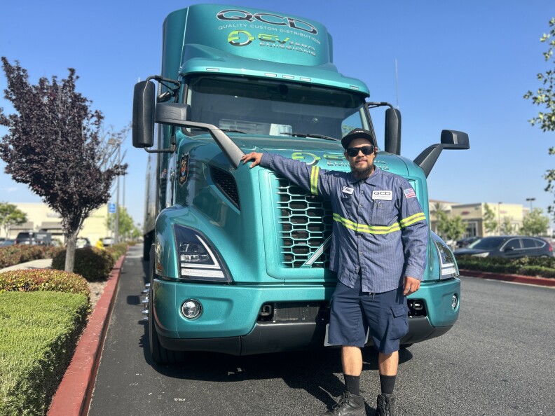 A man wearing a black baseball cap, work shirt and blue cargo shorts wearing sunglasses stands in front of a teal big rig truck.