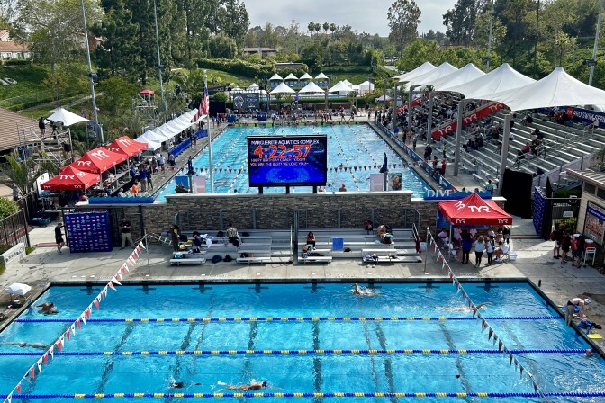 Looking down from above on two competition swimming pools. The one in the back has covered bleachers along the side and a large score board at the near end. 