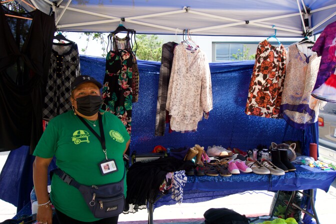 a woman wearing a face mask stands under a pop-up tent in front of a table laden with secondhand clothes and shoes.