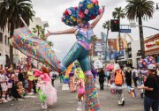 A wide shot of a person with a light skin tone standing on stills in the road in front of a crowd and Highland Avenue streeet sign. They have a bouquet of blue, yellow and pink flowers for hair and are wearing a bright blue and colurful outfit. Their arms are raised in the air while one leg is kicked up.