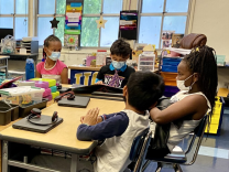 Four elementary school students, all wearing face masks, sit around a table in a classroom crowded with desks, bins of supplies and wall art.