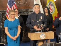 A Black woman in a blue dress and glasses folds her hands while listening to an Asian man in a police uniform address a crowd.