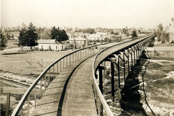 A black and white view of the California Cyclceway looking north from Raymond Hotel. The Santa Fe tracks are below and Fair Oaks Avenue is on left side. The cycleway is a wooden elevated path that curves as it extends away from the camera.