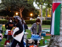 A young person wearing a face mask carries a crate of bottled water as others set up camp on a lawn.