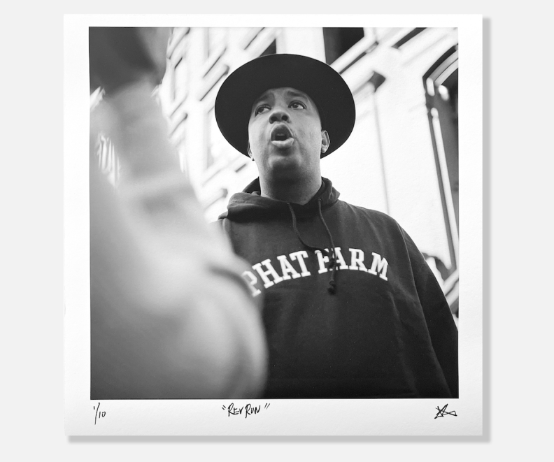 A black-and-white photo of Rev Run wearing a hat and a Phat Farm hoodie. 