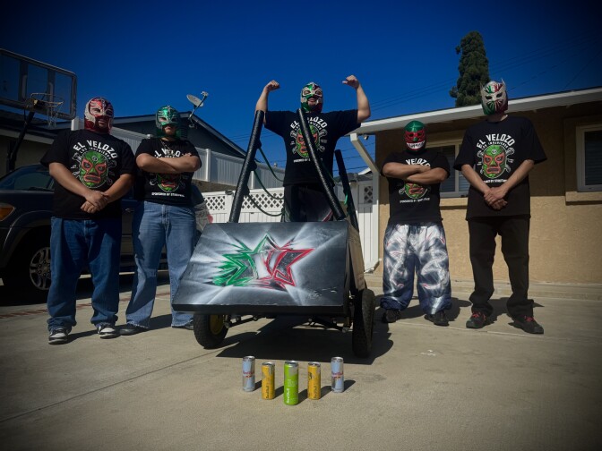 A spray-painted silver soapbox car with red, white and green accents. Five people in Lucha libre masks and matching black shirts are posing around the car, with one person standing in the driver seat with both arms raised in the air to show off muscles.