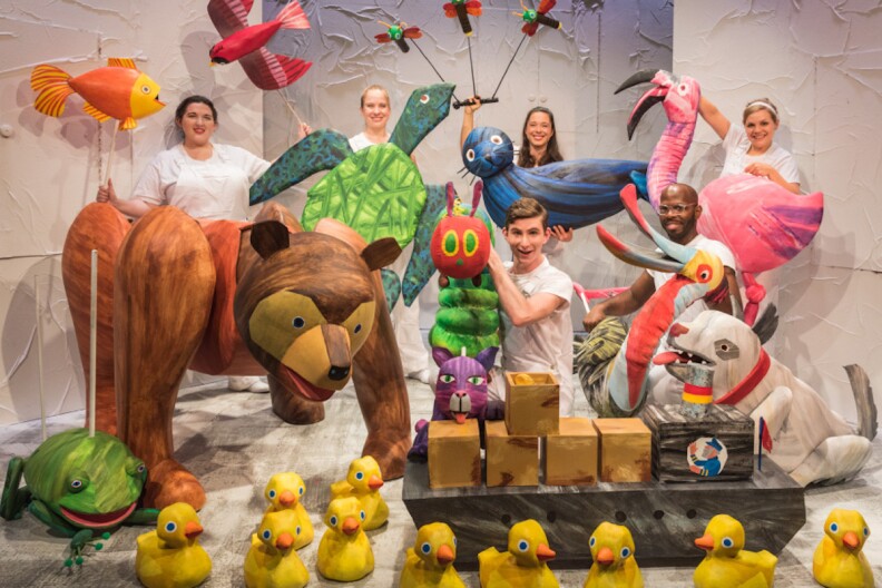 Five puppeteers stand with characters from the 'Very Hungry Caterpillar' show. There are ducks, a flamingo, fish, birds, a dog, a frog, and the titular caterpillar.