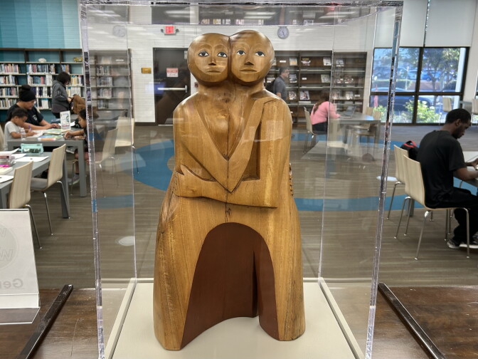 A wood sculpture of two people embracing, made from one piece of wood. The faces are turned towards the viewer.