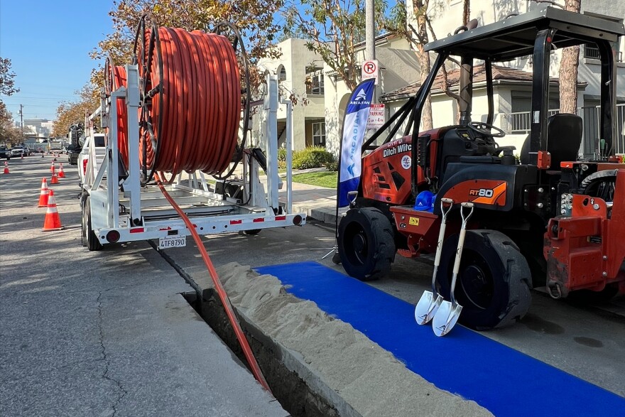 A wide view of the outdoors, showing a large spool of orange cables with the cable going into a trench in the street. On the right is a small construction vehicle with two shovels leaning on it.