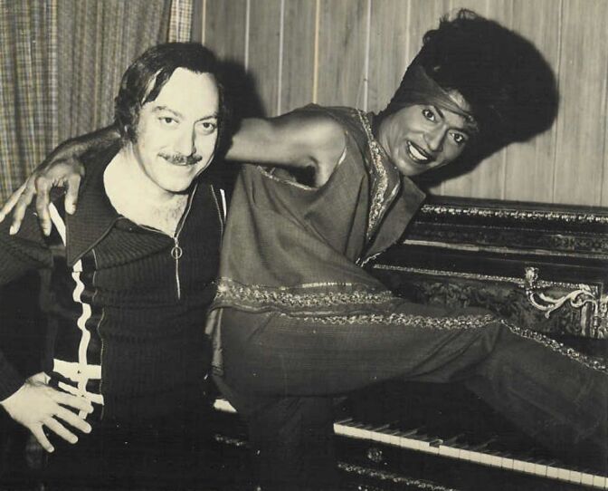 A man with a mustache has his arm around a man with his leg raised onto a puance and a tall Afro.