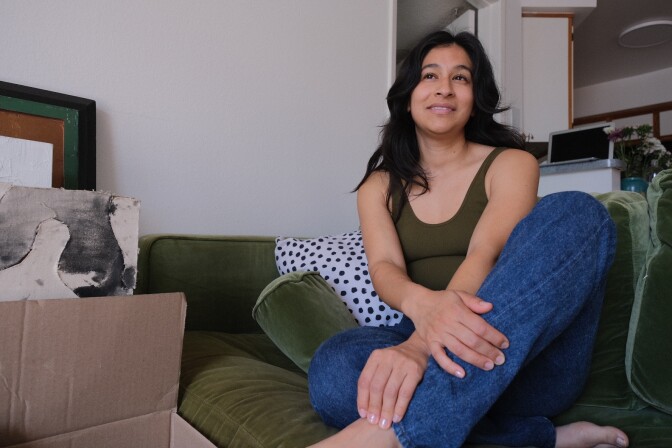 A woman with medium-tone skin sits cross-legged on a couch with boxes and artwork stacked nearby.