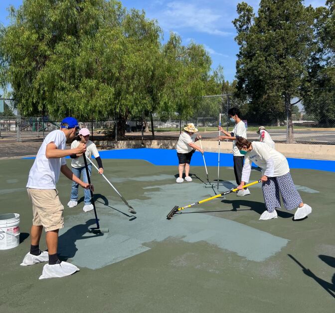 Half a dozen people are painting asphalt at a park using long paint rollers.