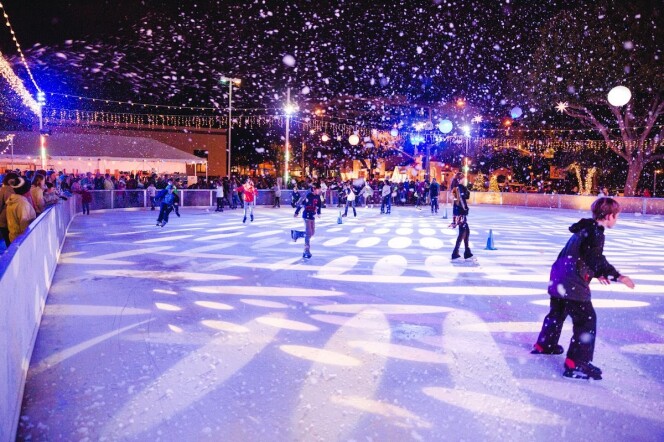 Snow falls over an ice rink at night with lights shining on skaters.