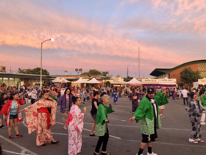 A line of Asian American and multiracial people, some in yukata and happi coats, dancing in a large open lot as the sky behinds them turns shades of pink and orange.