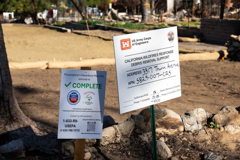 Two signs posted on a patch of dirt one reads "This property's Hazardous Materials removal is COMPLETE" and "California Wildfires Response Debris Removal Support" which lists an address and pin number.