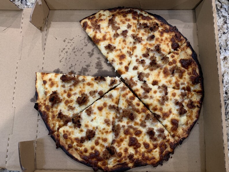 A pizza sits in a pizza box, with one slice missing. The pie is charred and cooked to golden brown. It is covered in alfredo sauce, and topped off with sausages, too. 