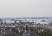 A view from above shows tract homes in Fontana, California, in the foreground and rows of white warehouse behind them.