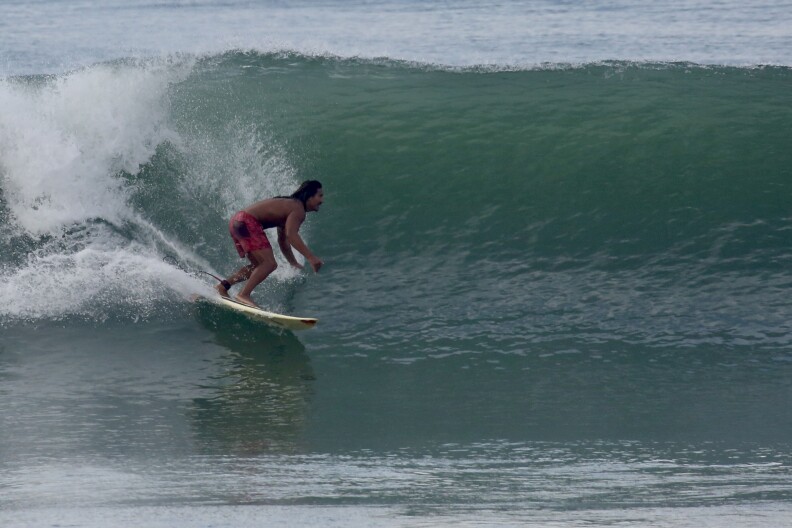 A man with long dark hear wearing red trunks surfs a big wave, with his back to the camera.