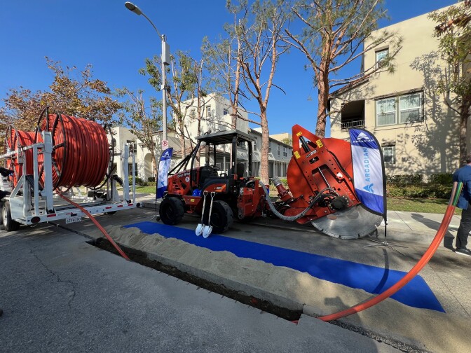 A red and black tractor is parked next to a giant spool of orange cable along a sidewalk in Boyle Heights. There are white residential buildings, green grass, trees in their winter state and a blue sky in the background.