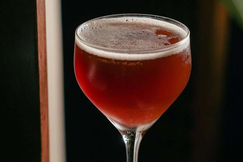 A small, stemmed glass hold a red-brown cocktail on a wooden surface.