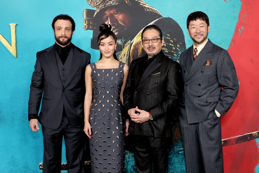 4 people stand behind a promotional backdrop for the Hulu series Shogun. The farthest to the left, a man wearing a three-piece, matte, all black suit; he's also got short, curly hair and full beard. Center left is a woman with a gray dress that has stylized holes throughout the dress; she has long hair that runs down her back but is also partially held up in a bun. Center right is a man dressed in a black, 3-piece suit that has glittery gray, vertical streaks; he's wearing a watch on his left wrist and is wearing glasses. Fourth person is a man with short hair and beard; he's wearing a great suit, white dress shirt and brown tie.