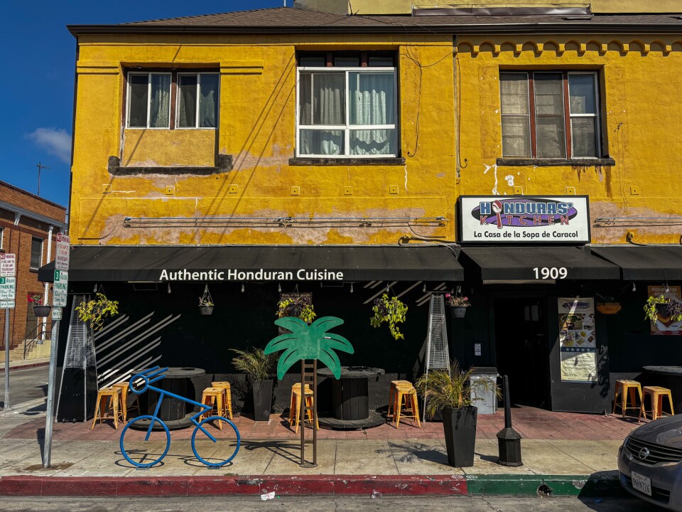 The bright yellow facade of Honduras Kitchen in Long Beach, with a black awning reading “Authentic Honduran Cuisine.” Outdoor tables and stools line the sidewalk beneath hanging plants and a palm tree cutout.