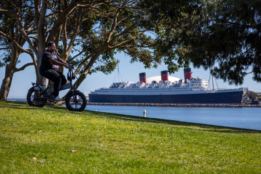 A man with dark skin wearing a black graphic t-shirt, black pants and white shoes rides an electric bike along a grassy path. A large black and white ship with three red and black smokestacks can be seen on the water in the background.