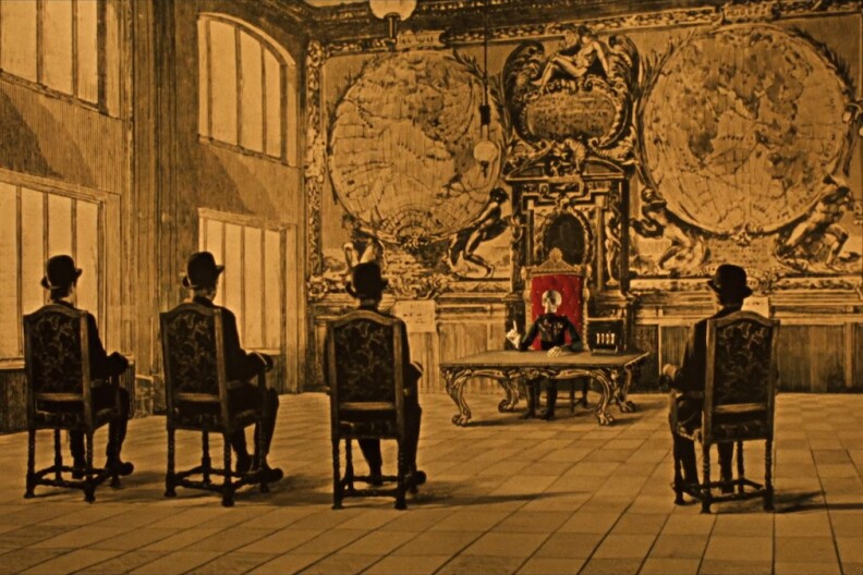 Four men in suits and hats sit with their backs to us while a skeleton in a red chair addresses them in a drawing room fit for king.