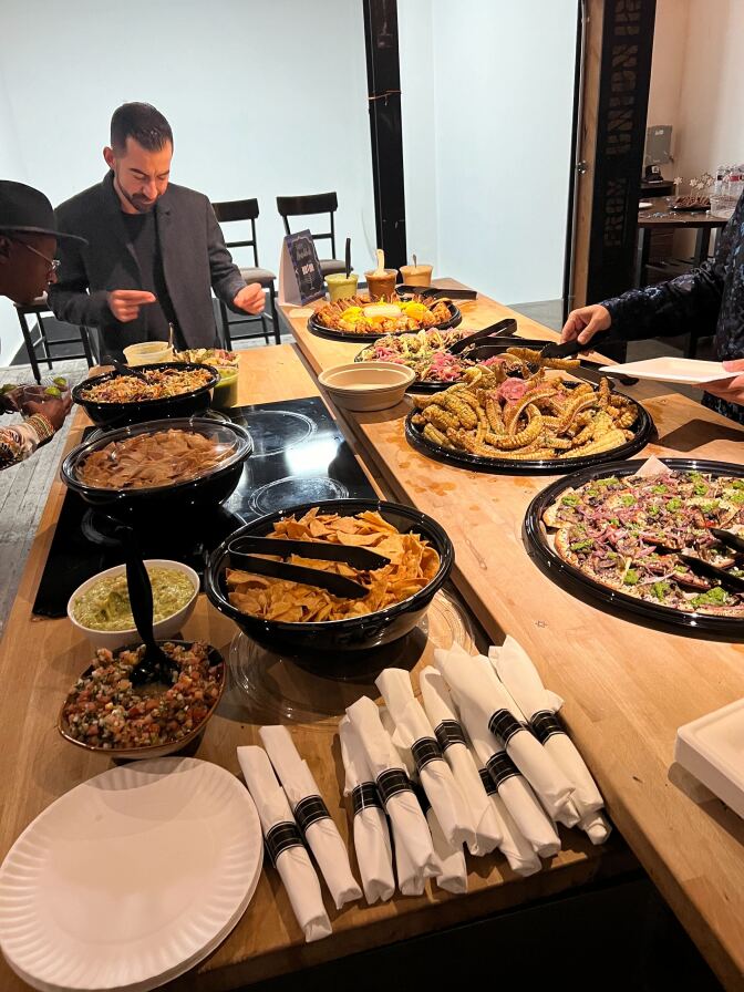 A man with facial hair wears a dark suit while bowing and gesturing over multiple plates of food at a table.