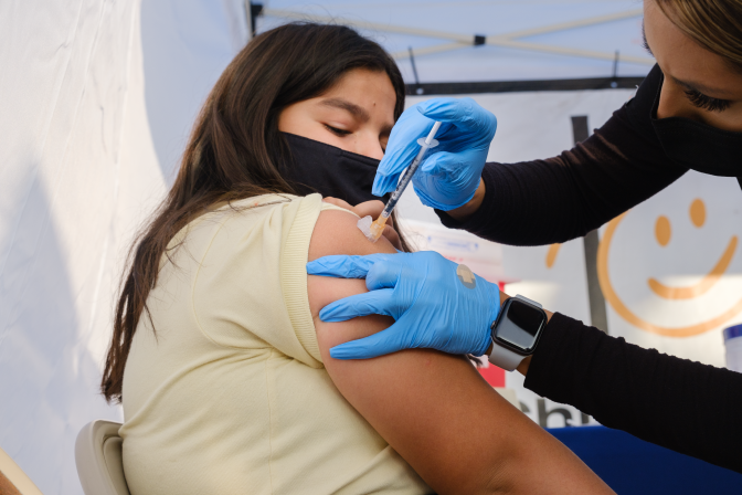 A girl gets the COVID vaccine on her right arm from a health expert. Both have their faces covered.
