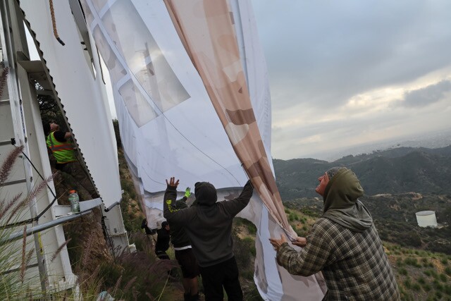 Workers, including several in hoods, seen holding the bottom of a large white and brown banner.