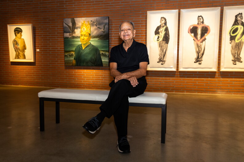 Cheech Marin sits, smiling, legs crossed, on a white bench in front of several pieces of art behind him. In one, Marin wears a crown, with seagulls flying behind him in front of a body of water. On the far left is a nude woman's back, the woman looking over her shoulder, a large tattoo on her back. On the right is a triptych with a person in front of illuminated letters saying "I [HEART] U."