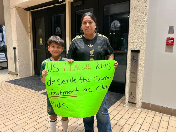 A boy wearinga. green soccer uniform and his mom who is wearing a black soccer jersey stand in City Hall holding a green sign that reads, "Us League kids deserve the same treatment as club kids."