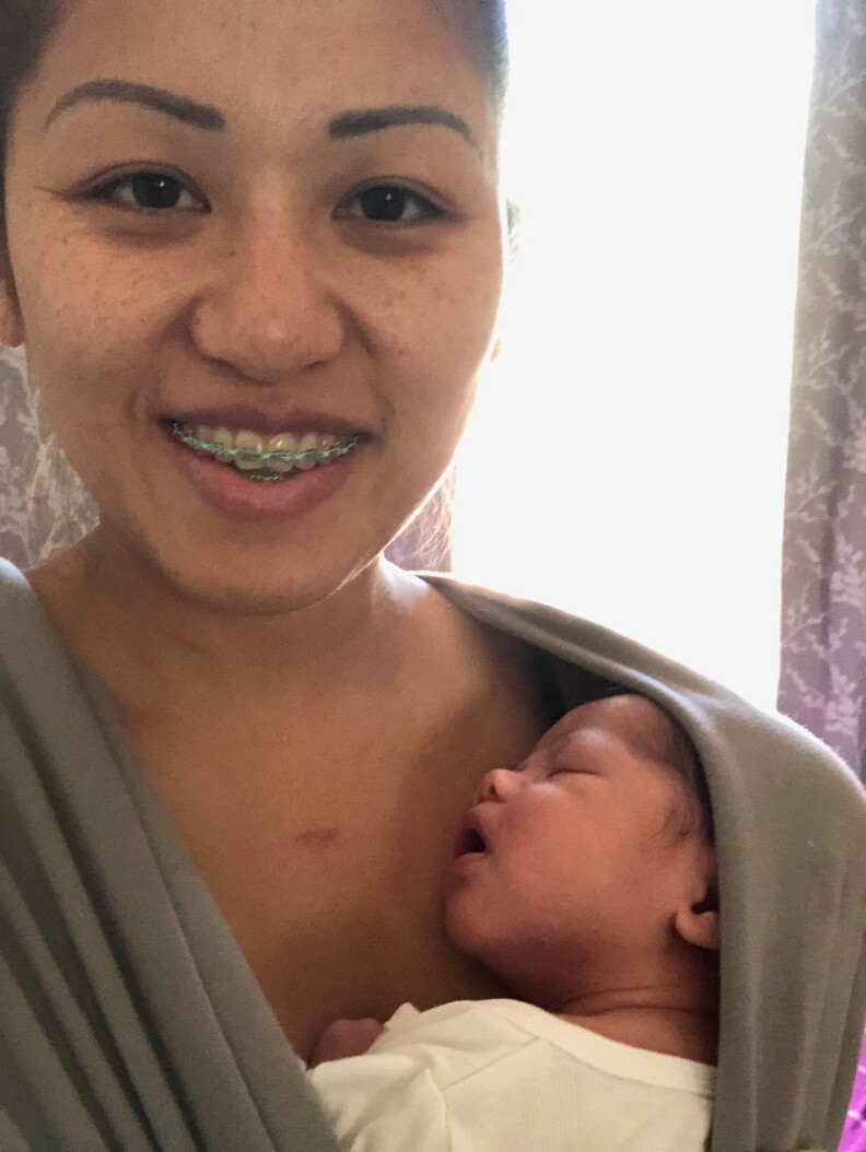 A Vietnamese woman with medium-toned skin and braces looks into the camera. Her sleeping newborn baby is resting his head against her chest, wrapped in a soft grey baby wrap carrier.