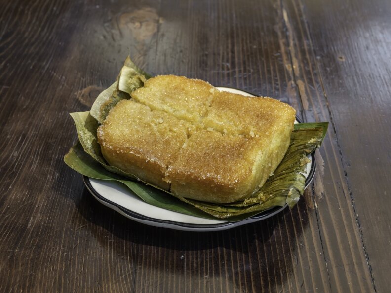 A beautiful rich-looking piece of cornbread is cut into four quarters. It sits on banana leaves on a white plate, on top of a brown table.