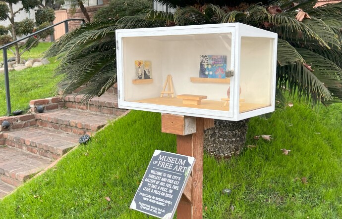 A white box with two transparent walls that showcase small works of art, including a ceramic nose and drawings. The box, located in a home's front yard, is accompanied with a sign that reads "Museum of Free Art"