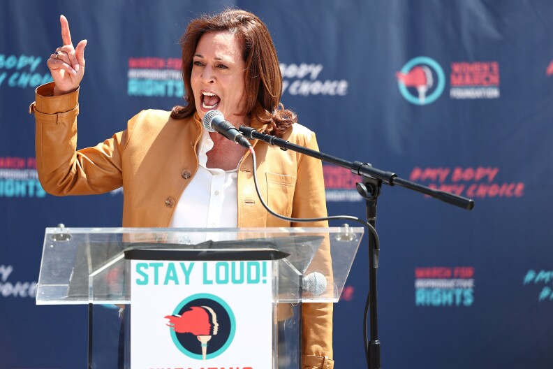 Awoman with dark tone skin speaks at a lectern. Signs read: "Stay Loud"