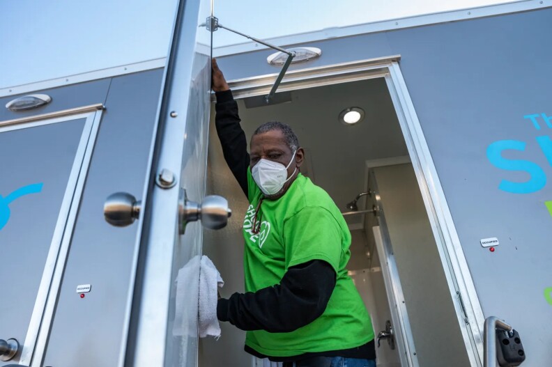 A Black man in a light green T-shirt, with short black hair that's starting to go gray, wipes the inside of the door of a silver Shower of Hope truck.