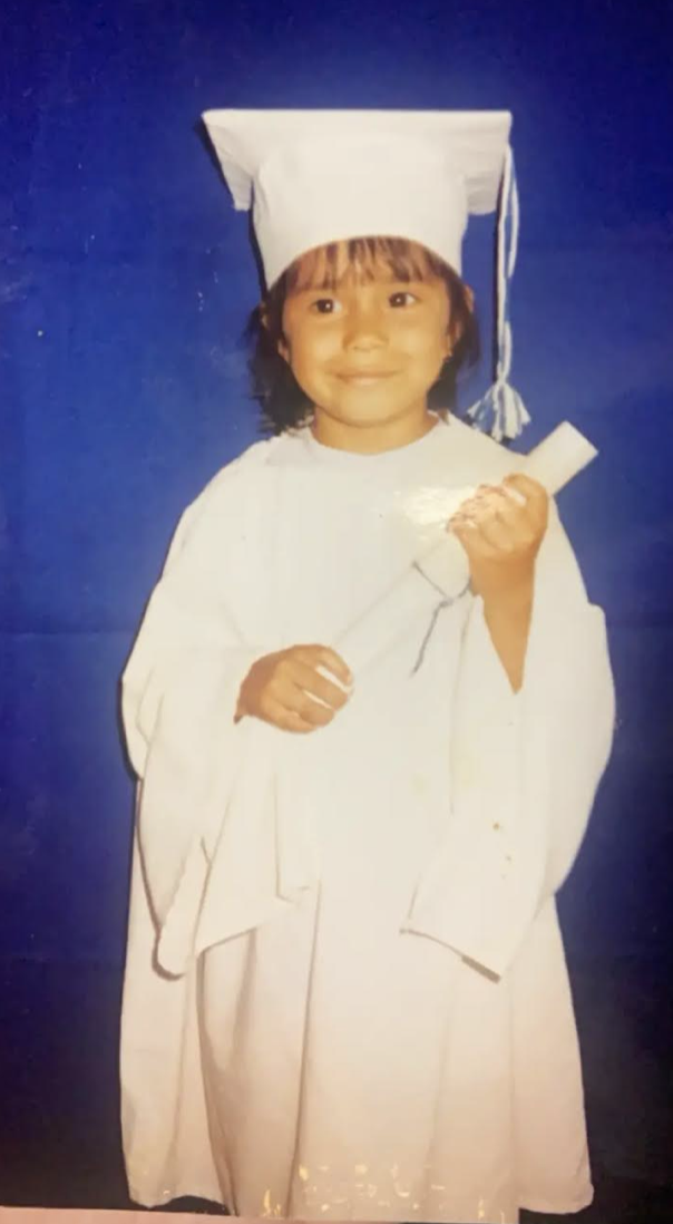 A young Flor wears white gown and graduation cap. She carries a rolled up diploma.