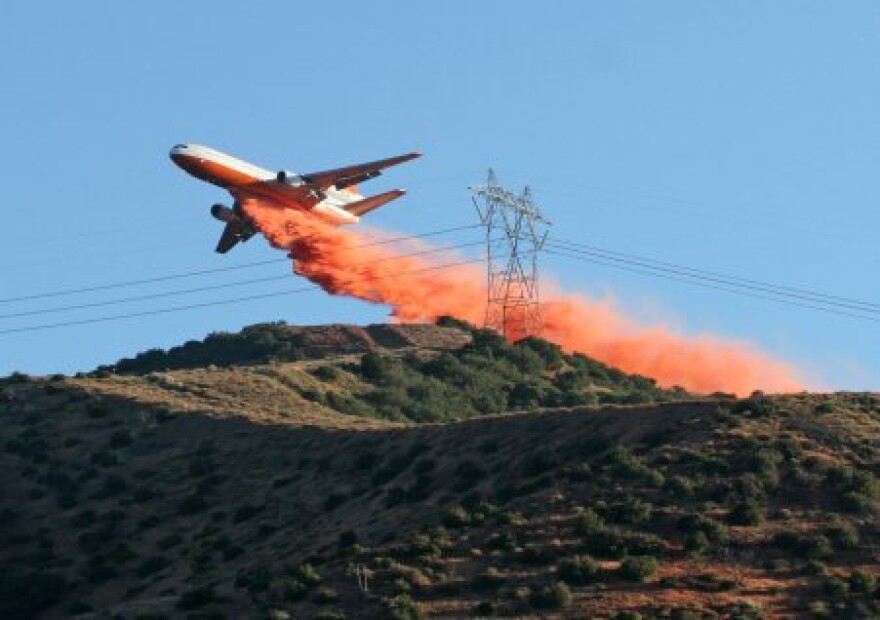 A large firefighting plane drops fire retardant on a mountain ridge near power lines. The retardant landing on insulators can cause a high power line to short out.