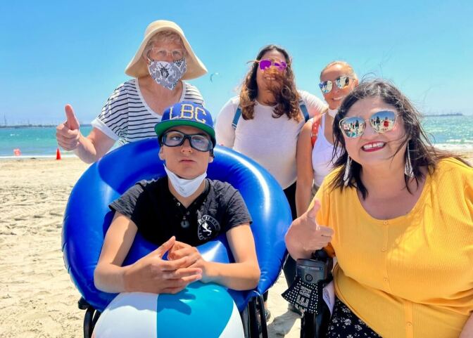 A person in a wheelchair at the beach. Four people are around him.