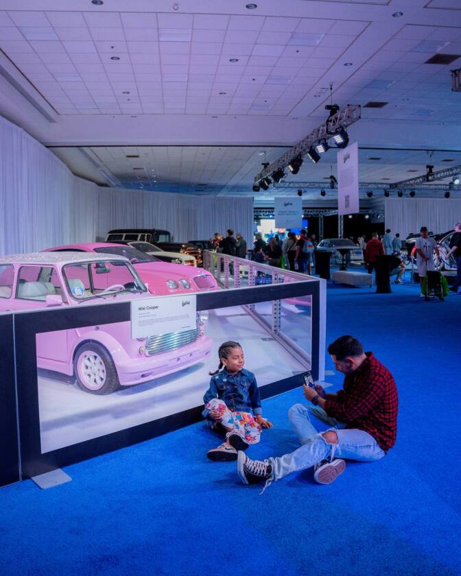 The ballroom floor at the Los Angeles Auto Show. An adult man takes a photo of a young girl posing in front a display of pink cars. They're both sitting down on the ballroom's blue carpet.