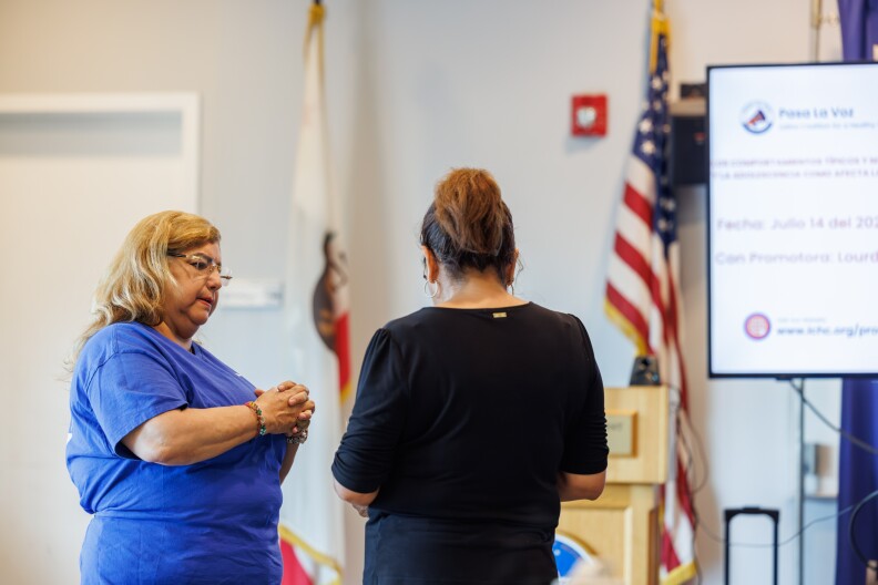 A woman wearing a blue tshirt is pictured speaking to another woman, shown from behind, wearing a black shirt. In the background is an American flag and a screen