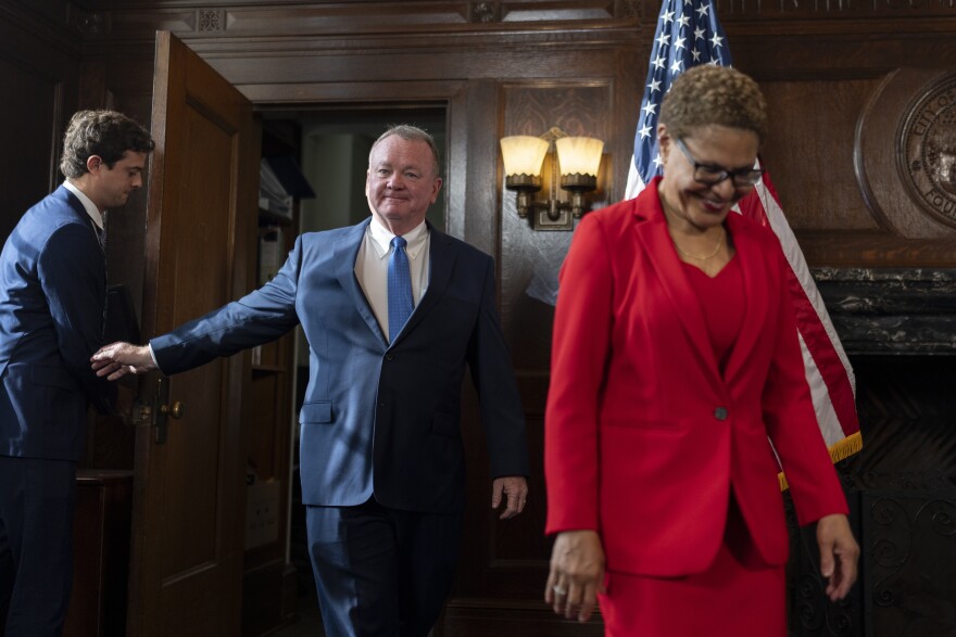 Jim McDonnell walks through a door held open by another man. He is in a blue suit and tie. He follows behind Mayor Karen Bass, a Black woman in a red suit jacket, shirt and skirt.